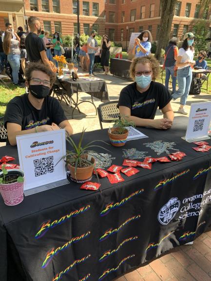 Ryan Auld and Abi Bickford smiling behind masks at the SCC table at the CAS Club Welcome Event. They are both wearing Rainbow Beaver shirts. The table has a black cloth, skittles, stickers, plants, and printed QR codes on it.